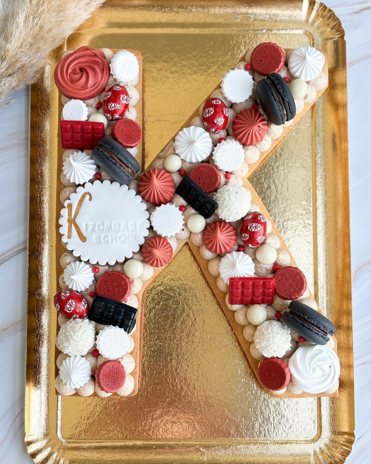 Letter K shaped cake decorated with red and white macarons and frosting swirls.