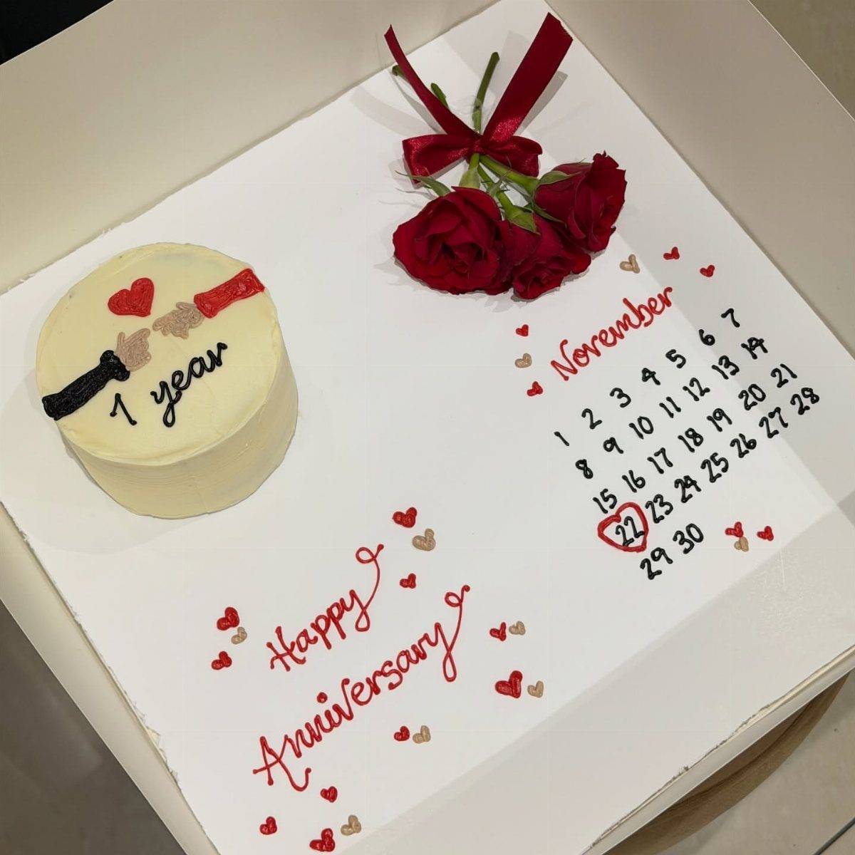 Small white anniversary cake with red roses and a calendar on a white board.