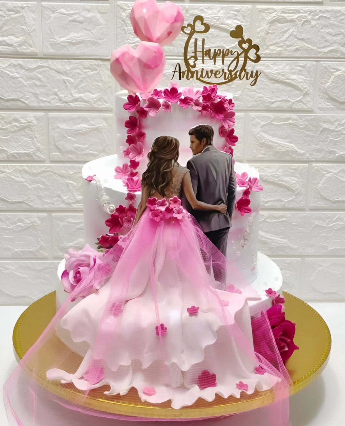 Three-tiered white anniversary cake with pink flowers and a couple figurine topper.
