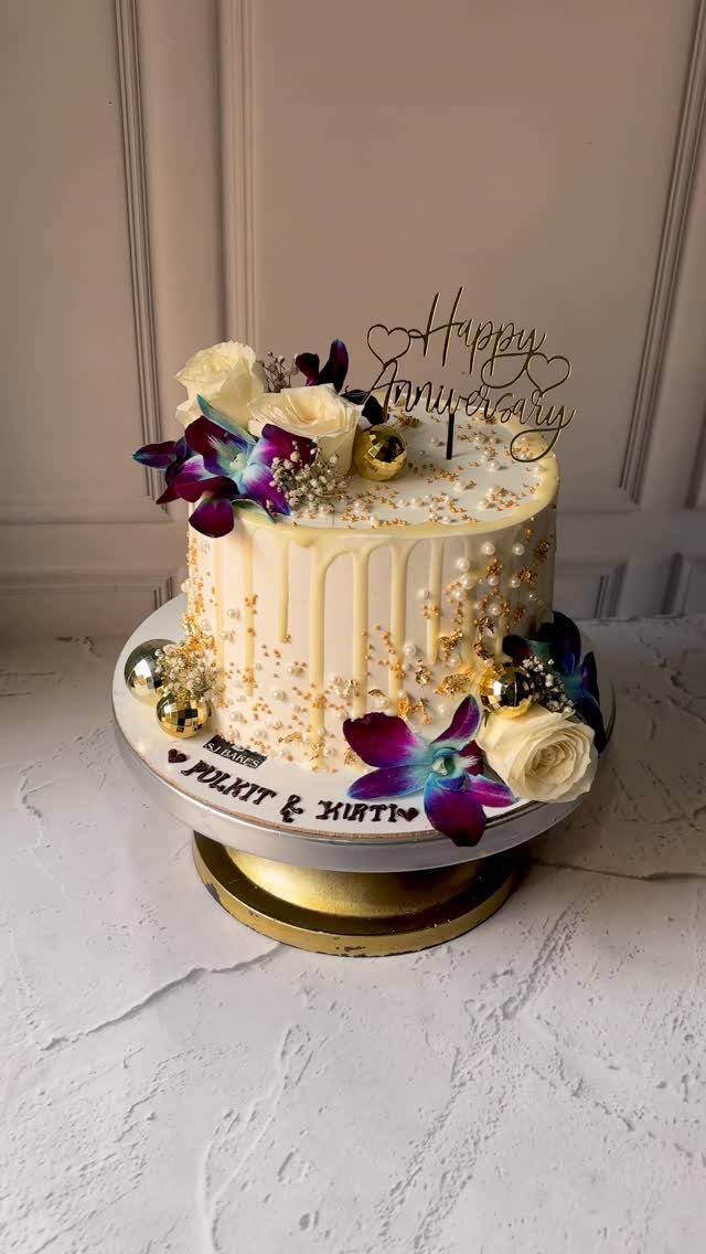 White anniversary cake with gold drip, white roses, purple orchids, and a topper.