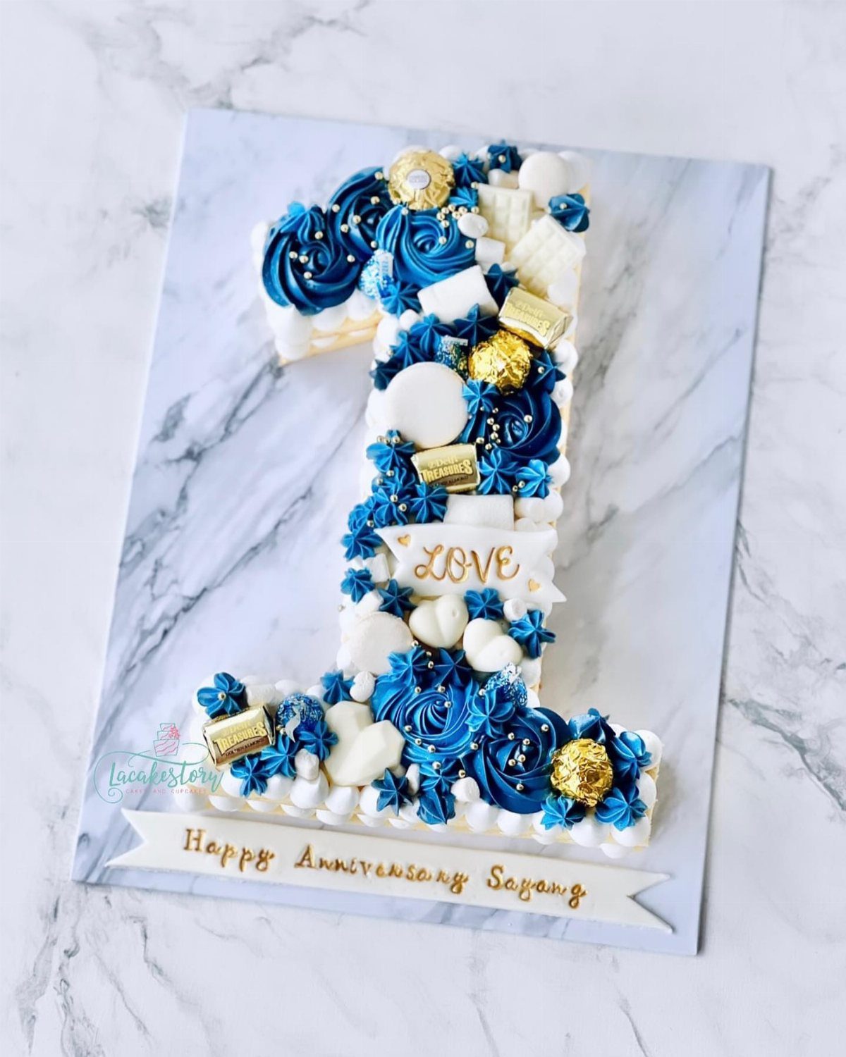 Number one shaped cake with blue and white frosting and gold chocolates