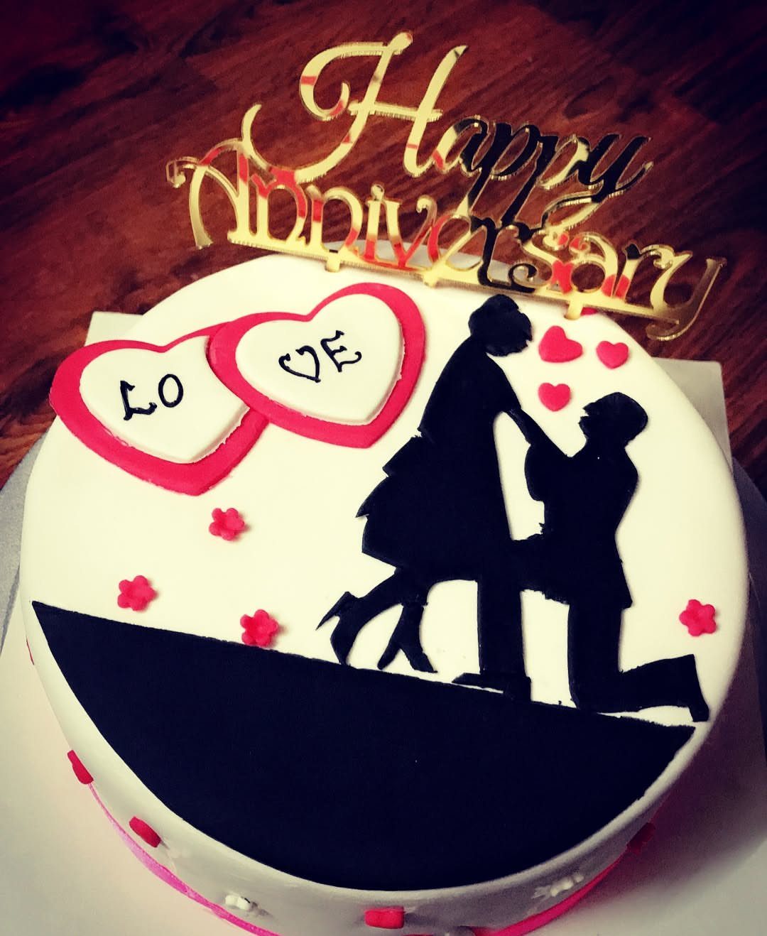 White anniversary cake with black couple silhouette, red hearts, and gold topper.