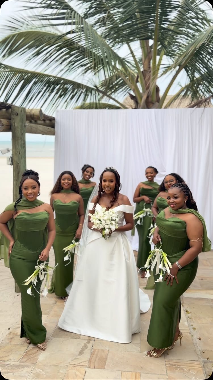 Bride in white gown posing with bridesmaids in olive green dresses on a beach.