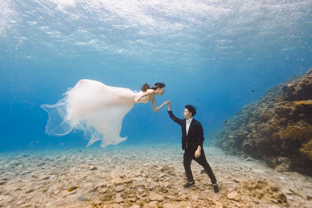 Couple in wedding attire holding hands while floating underwater in a clear blue ocean.