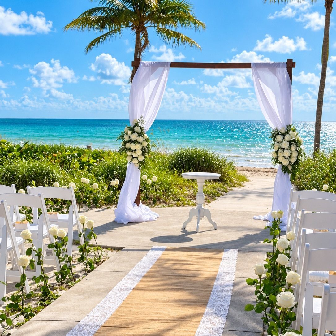 Wedding ceremony setup with white arbor, chairs, and aisle runner on a beach.