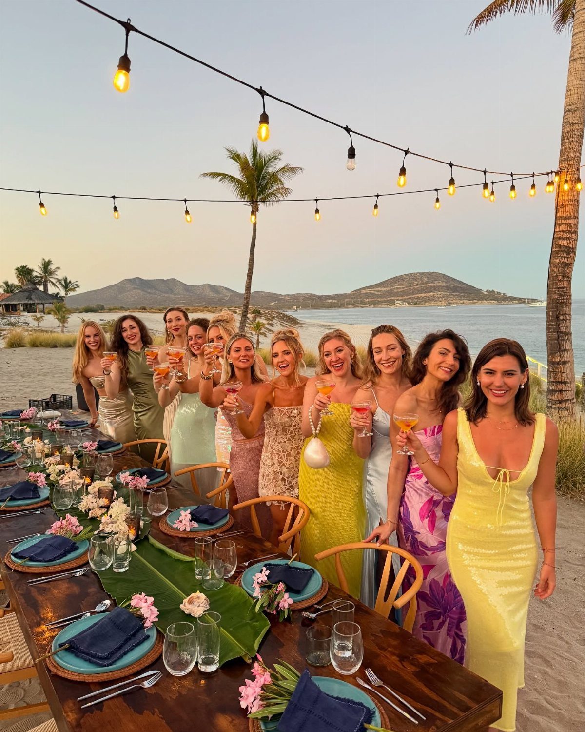 Group of women in colorful dresses smiling and holding champagne at beach dinner.