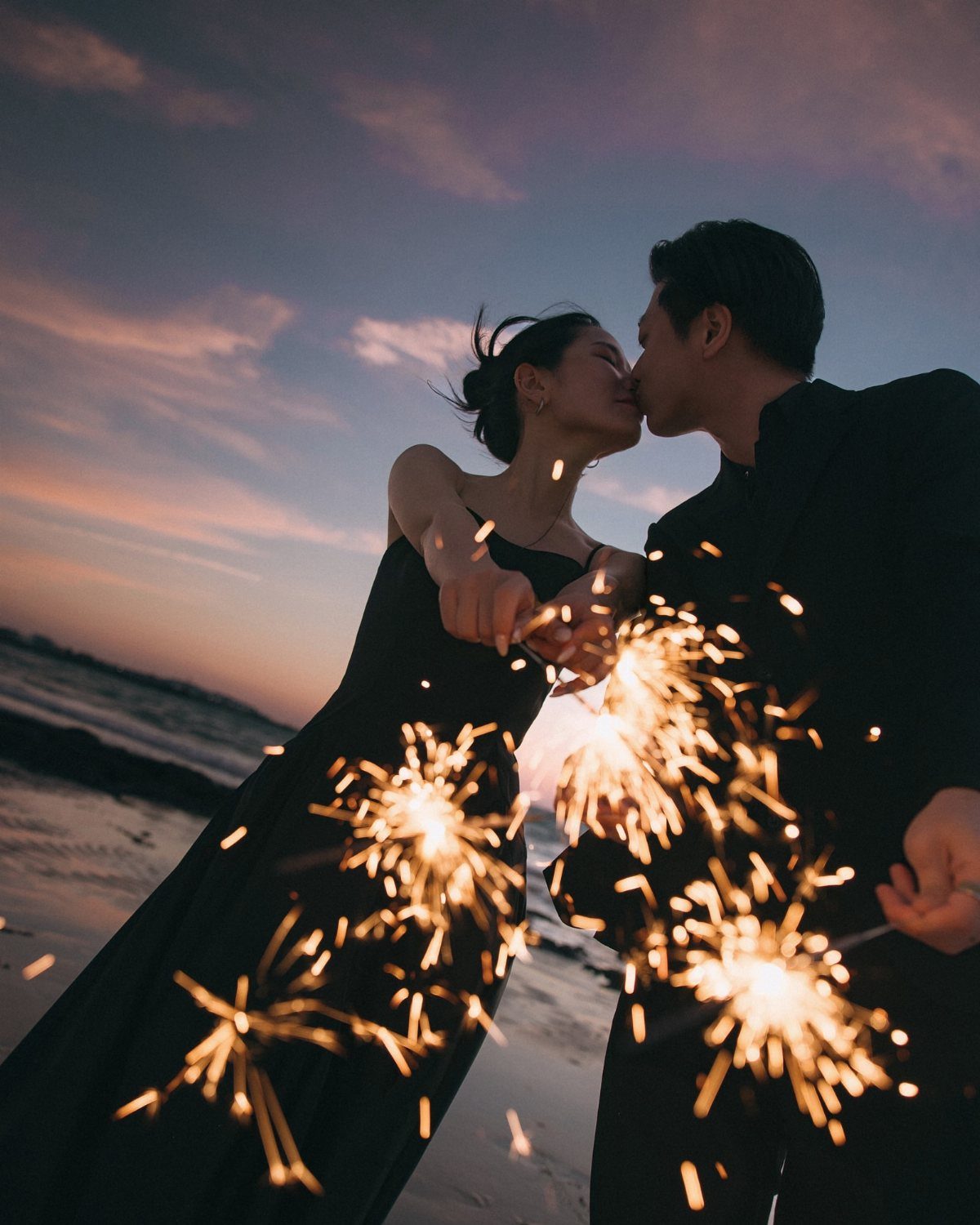 Couple in black formal wear kissing on a beach while holding lit sparklers.