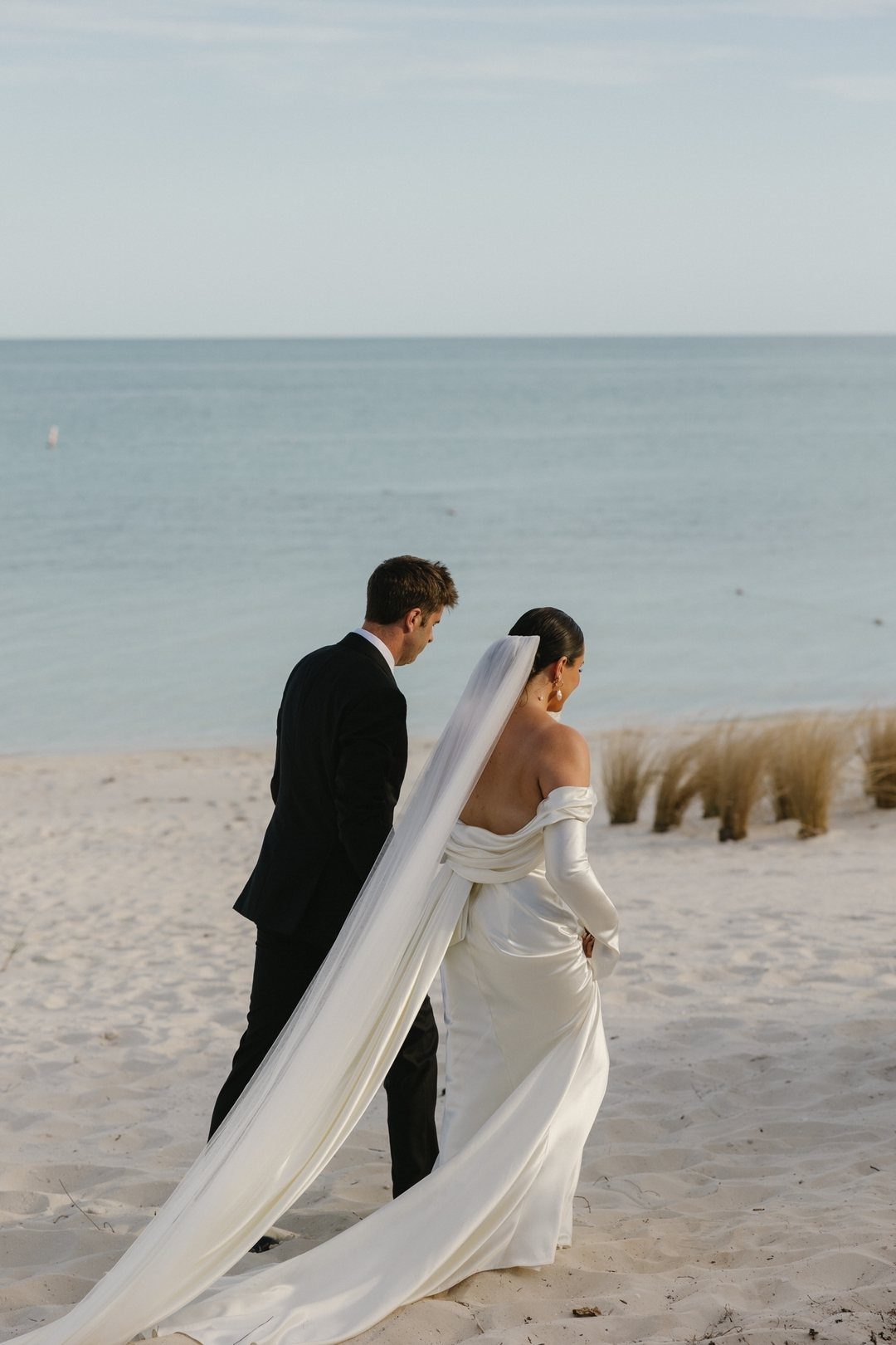 Bride and groom walking on a white sand beach toward the blue ocean.