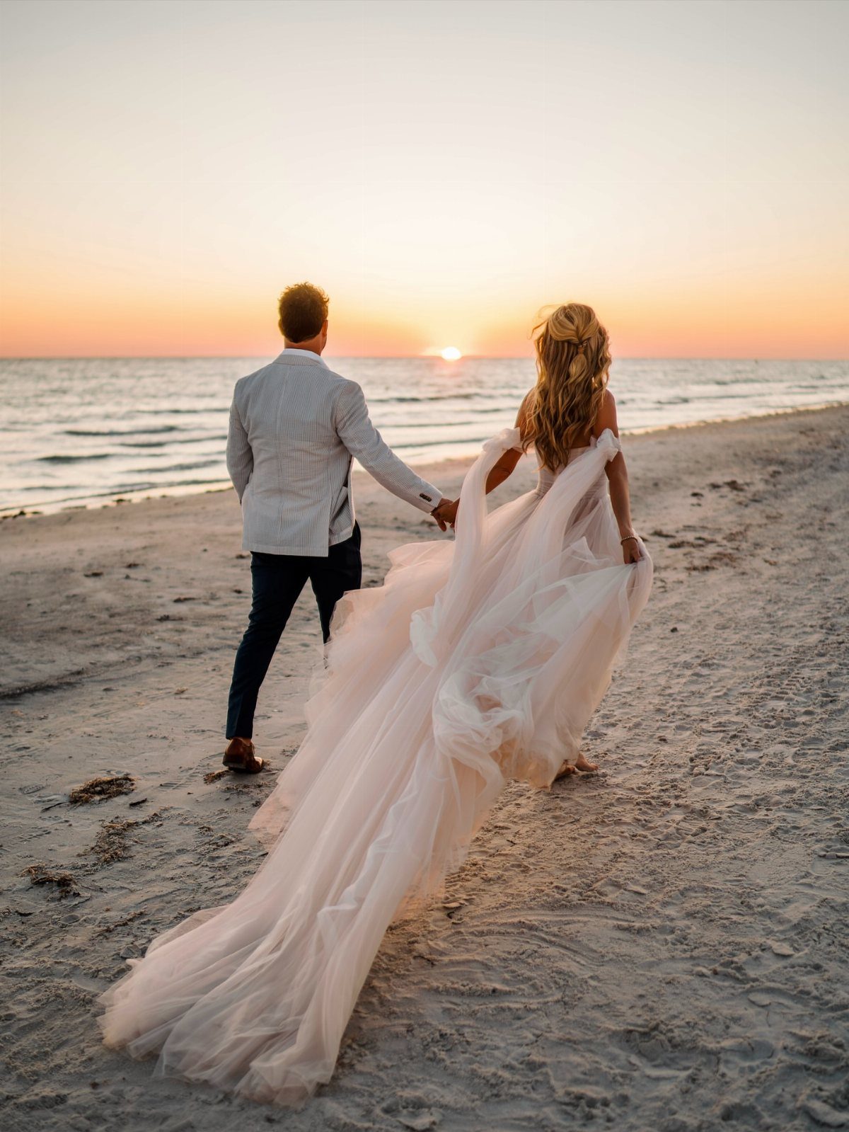 Bride and groom walking hand in hand on a beach at sunset.