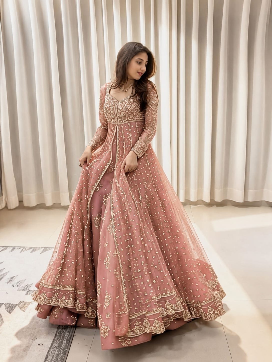 Woman in a dusty rose embroidered Anarkali suit posing against white curtains.