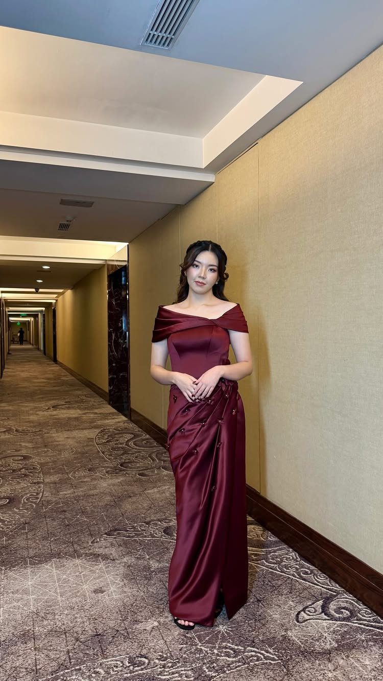 Woman in a burgundy off-shoulder satin evening gown posing in a hotel hallway.