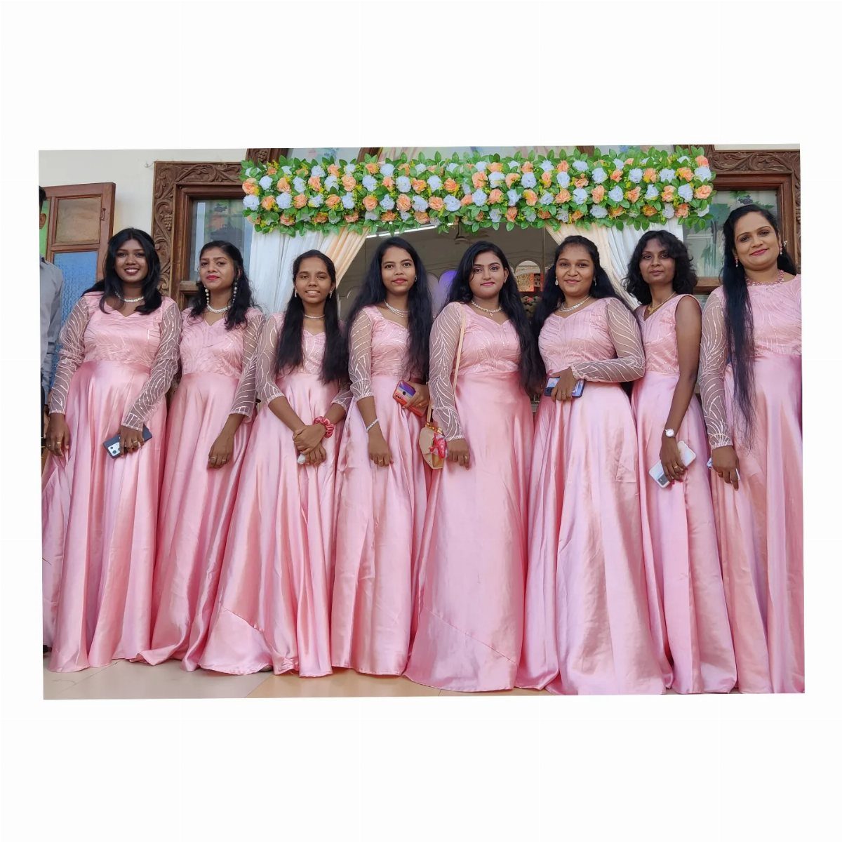Eight women posing in matching floor-length pink satin gowns with lace sleeves.
