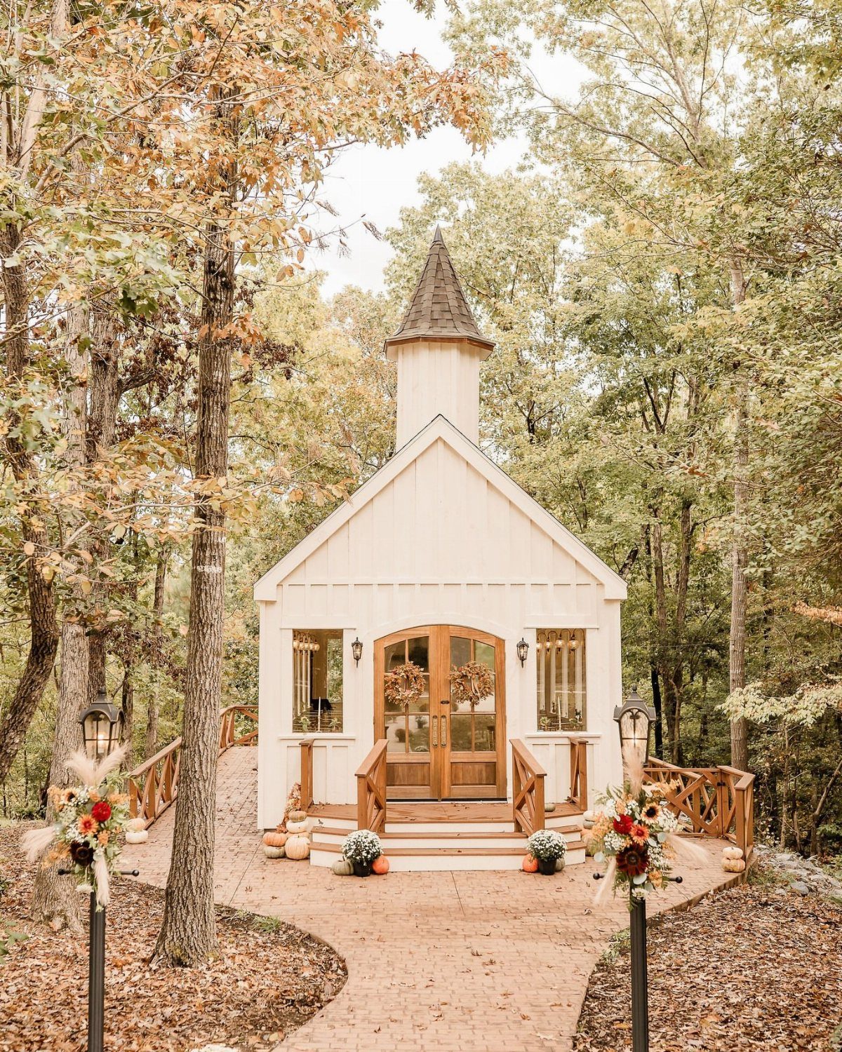 Small white wooden chapel with a steeple nestled in an autumn forest setting.