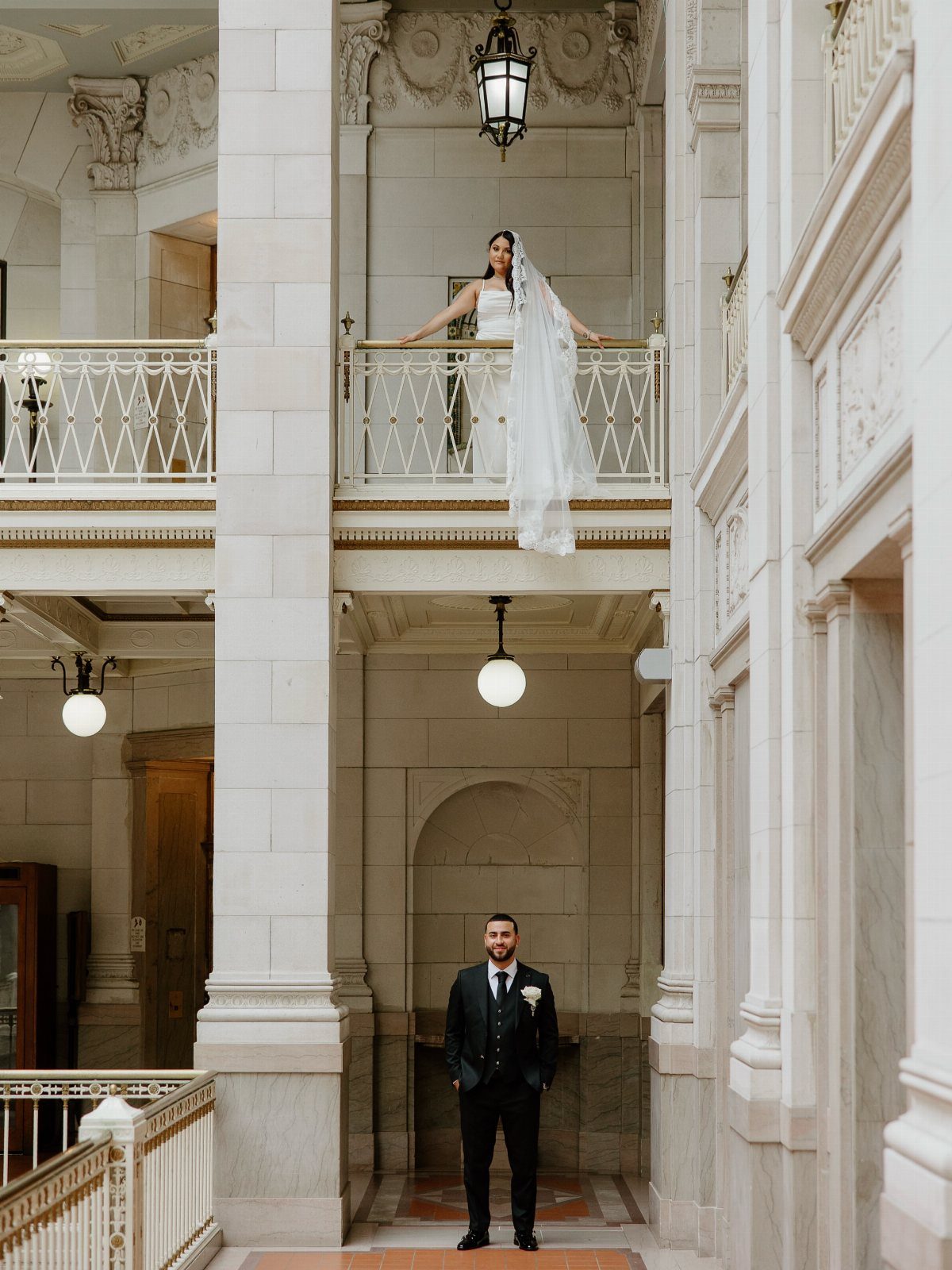 Bride on a balcony and groom below in a grand marble building interior.