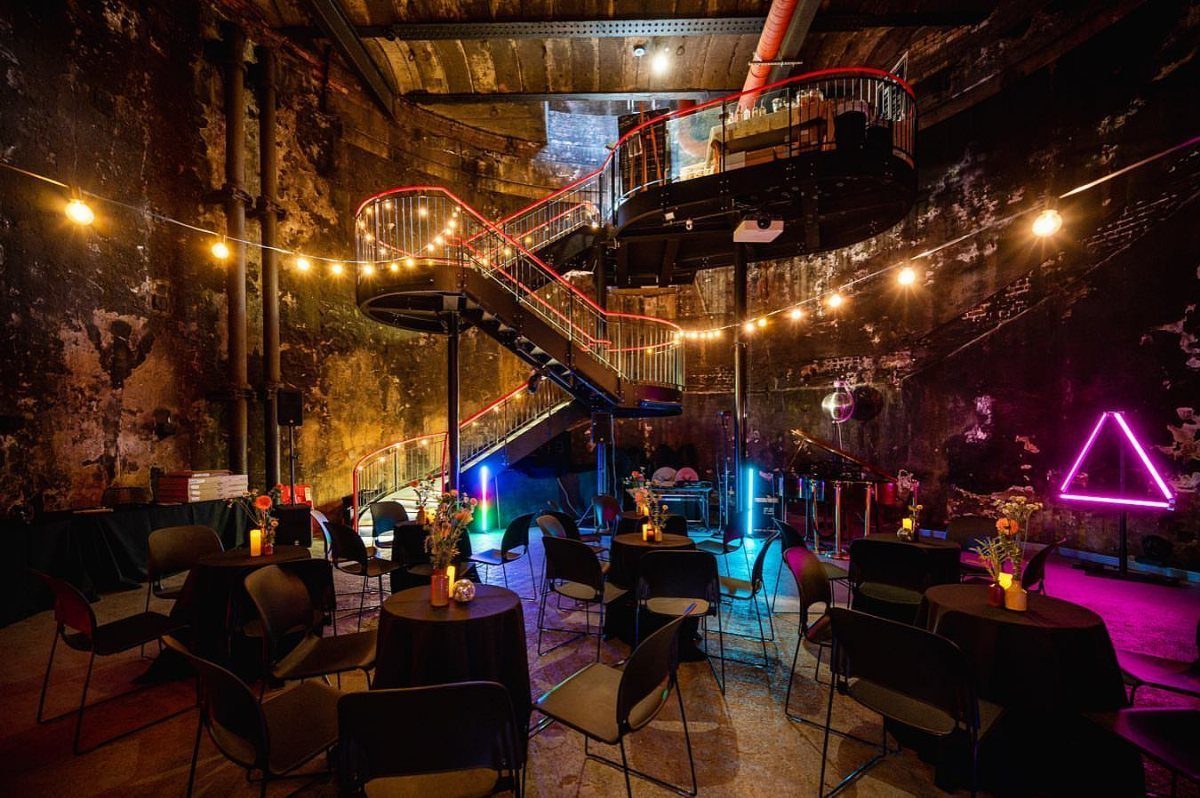 Industrial event space with black tables, string lights, and a red neon spiral staircase.