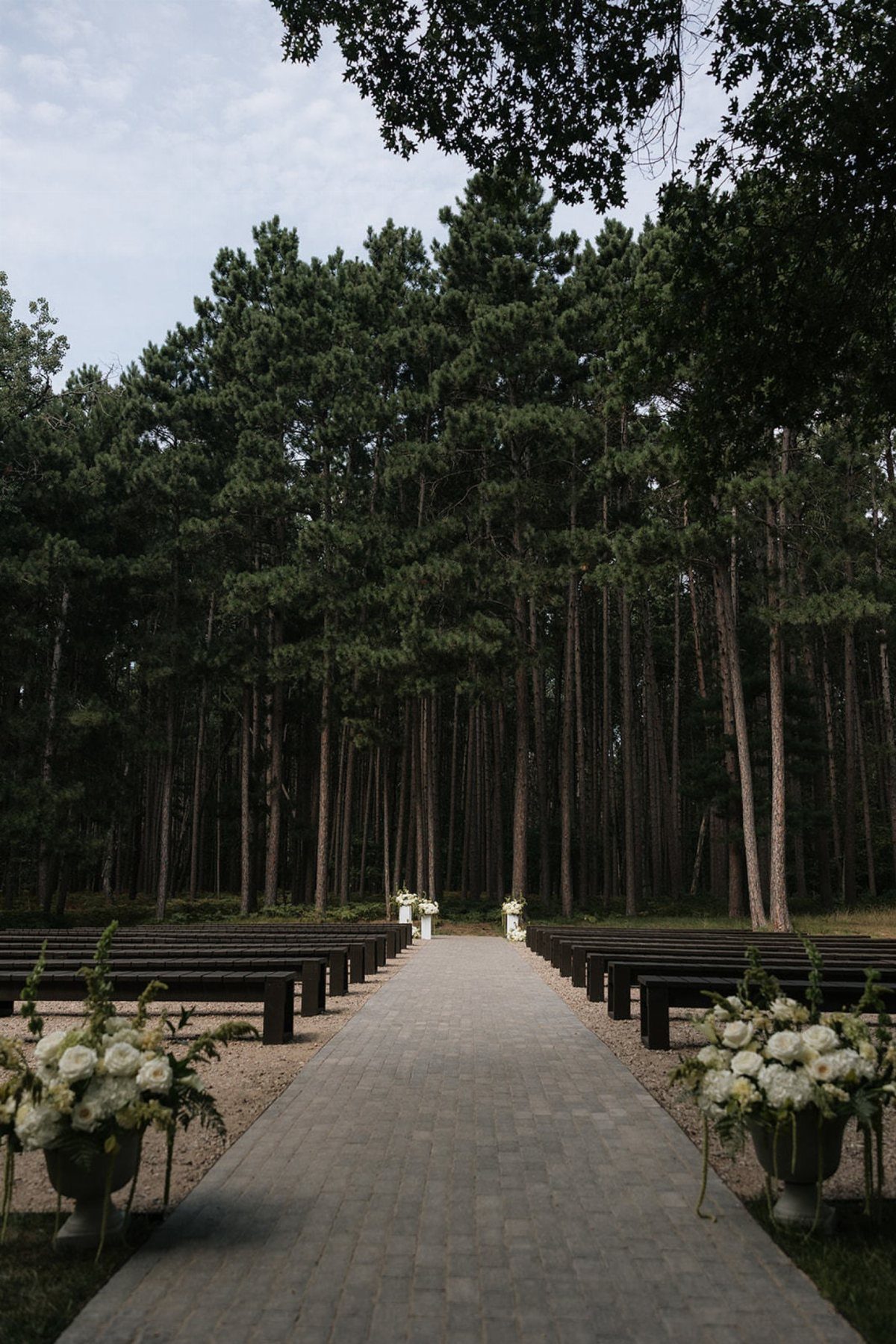Outdoor wedding ceremony aisle with wooden benches and white flowers before a pine forest.