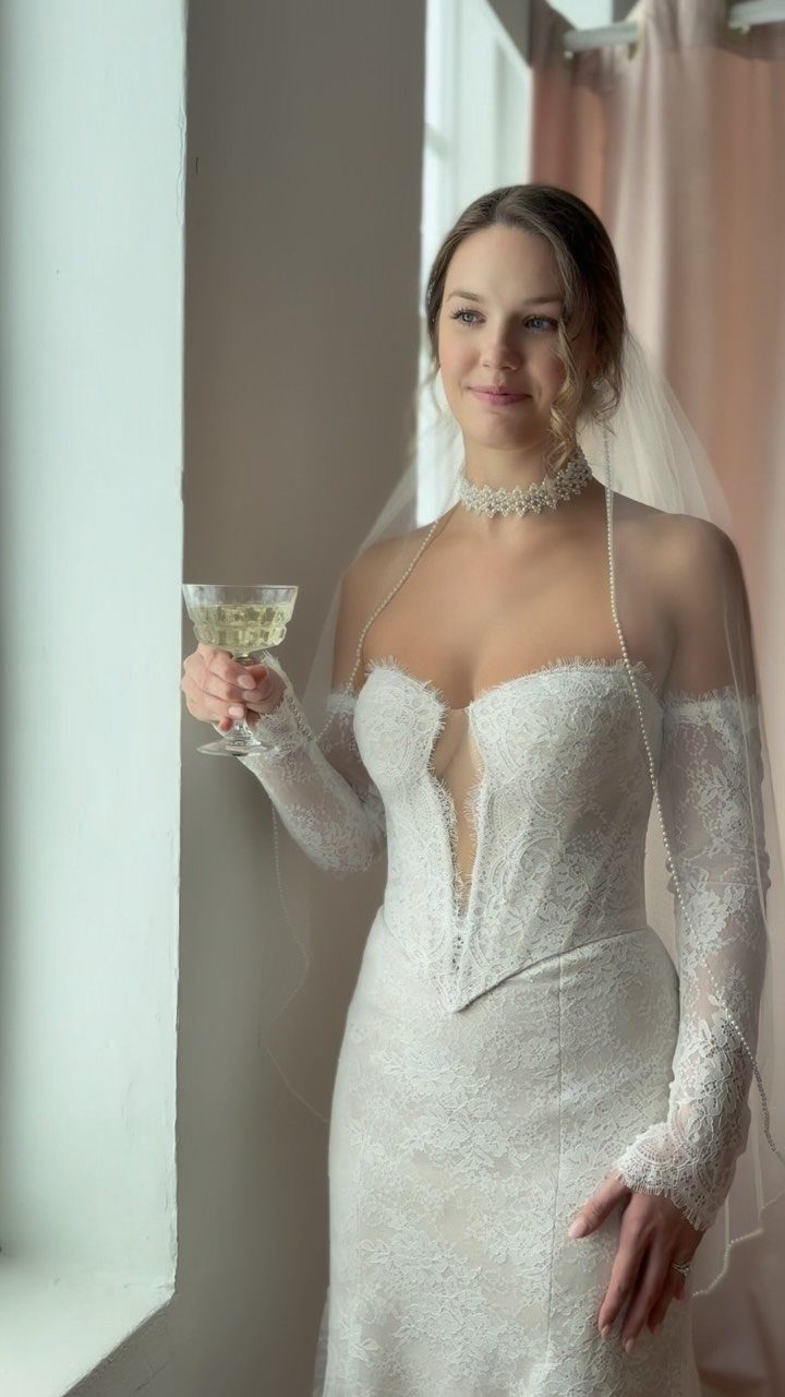 Claire Wedding Dress Styles: 11 Gowns to Inspire Your Look 10 Woman in a white lace wedding dress holding a glass of champagne by a window