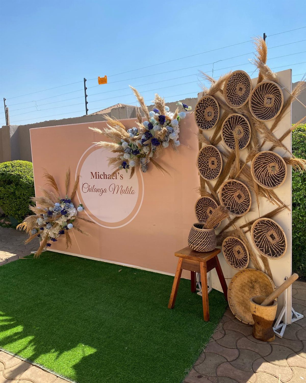 Photo booth backdrop with peach panel, blue flowers, and woven basket decorations outdoors.