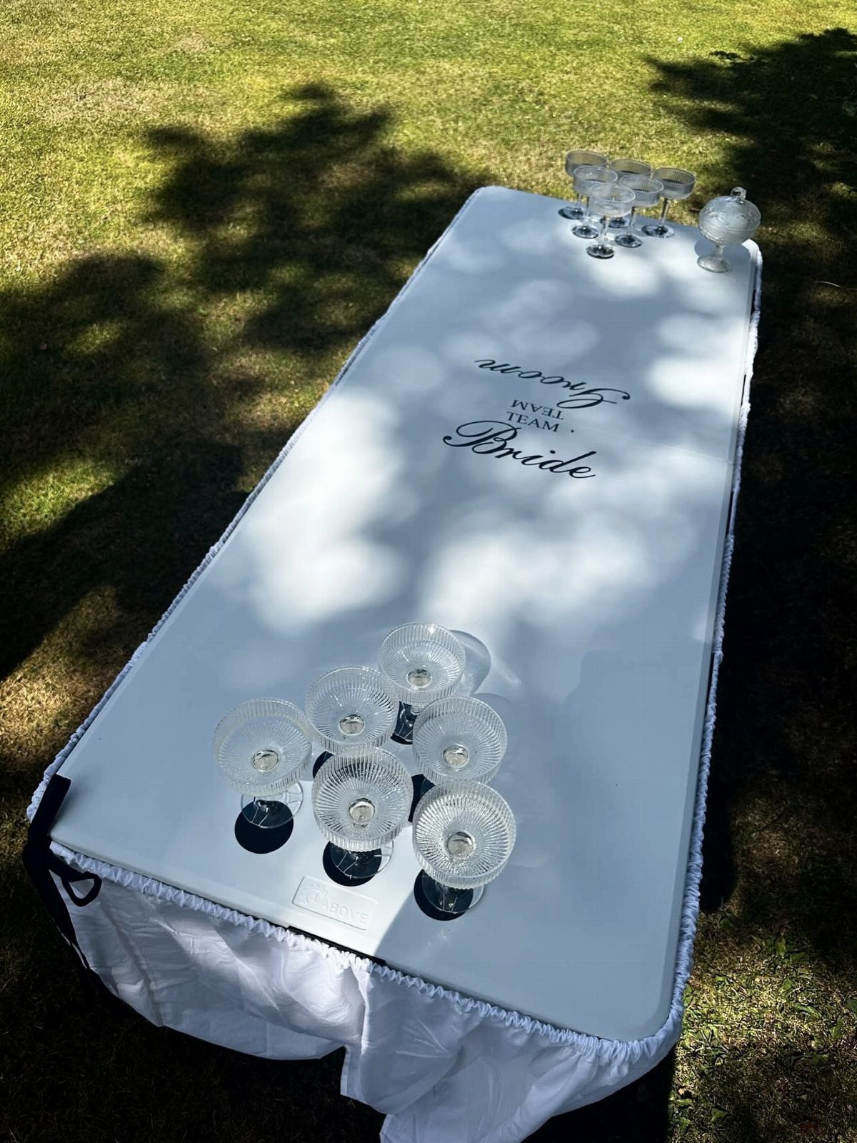 White prosecco pong table with champagne glasses on a sunny green lawn