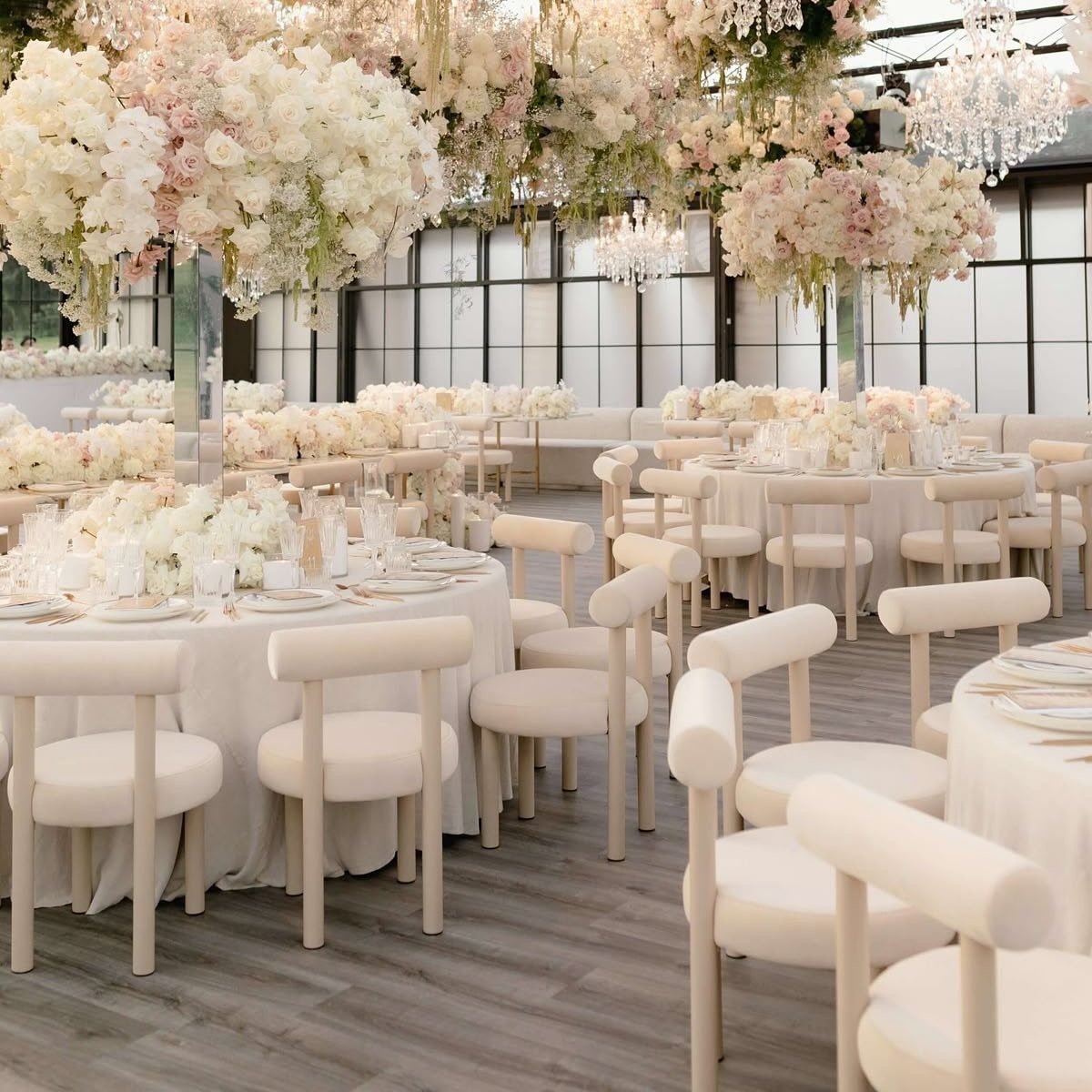 Cream-colored curved chairs around white wedding tables under hanging floral arrangements and chandeliers.