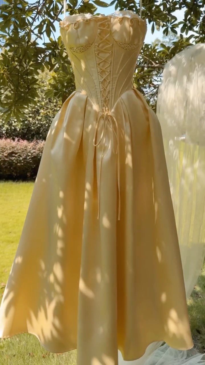 Gold satin ball gown with corset bodice hanging outdoors among green leaves.