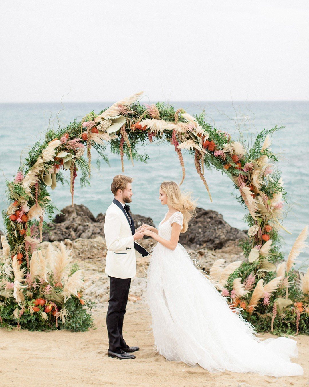 Bride and groom standing under a floral arch on a sandy beach by the sea