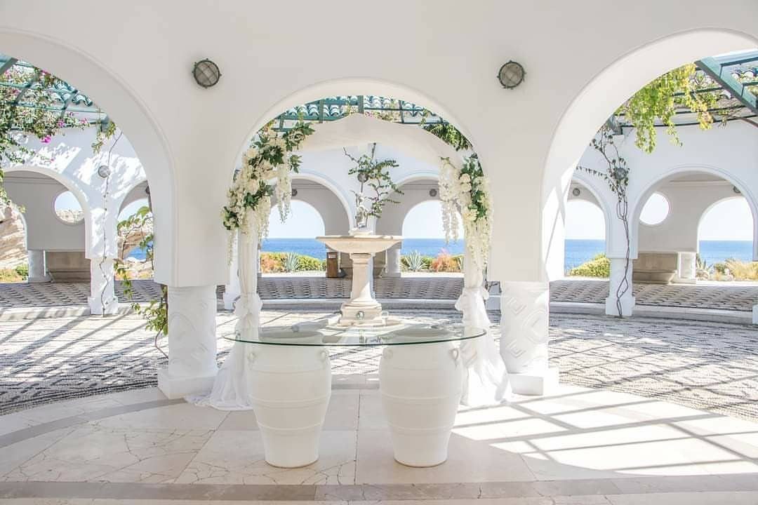 White arched pavilion decorated with flowers overlooking a blue sea for a wedding.