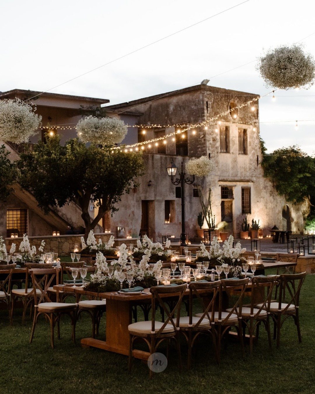Wedding reception tables on a lawn before a historic stone estate with string lights.