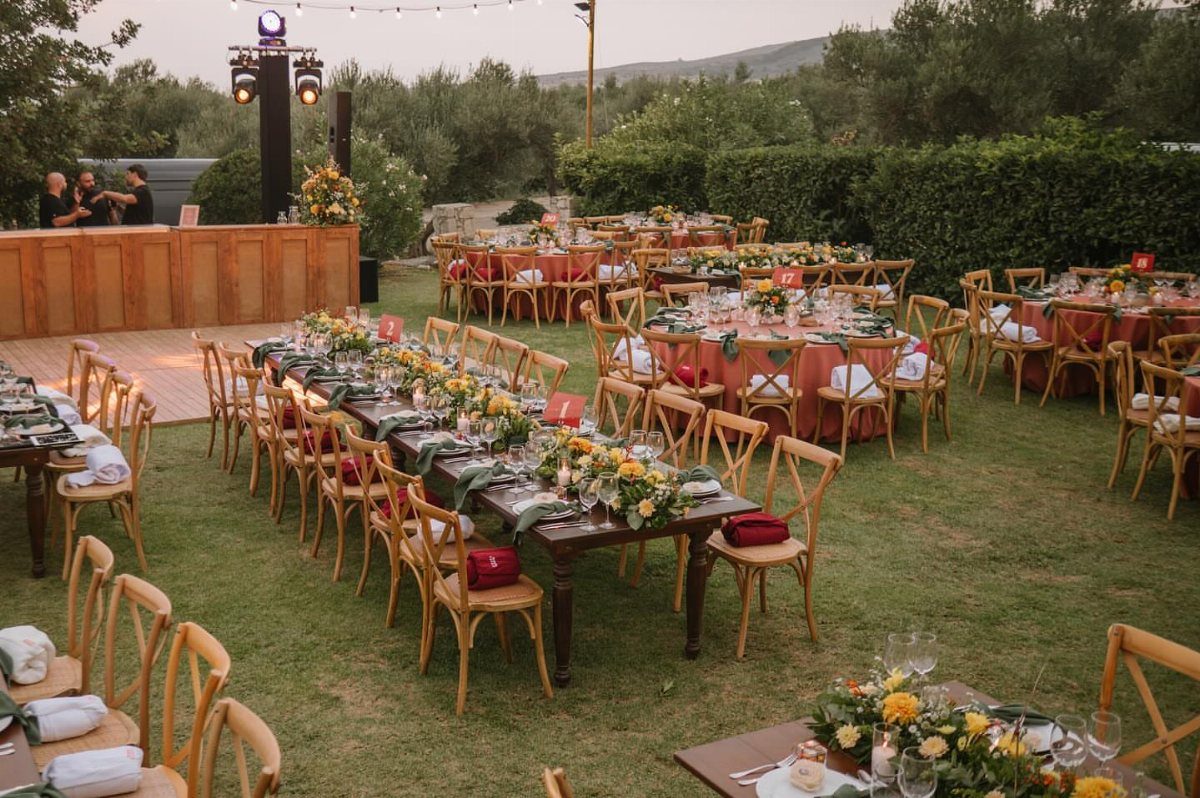 Outdoor wedding reception with decorated tables set on a lawn surrounded by olive trees.
