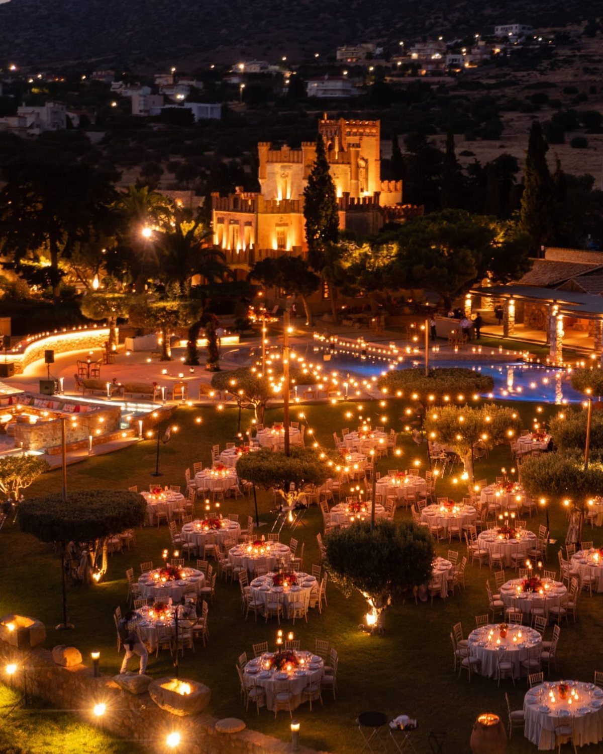Outdoor wedding reception with candlelit tables and string lights before a lit castle.