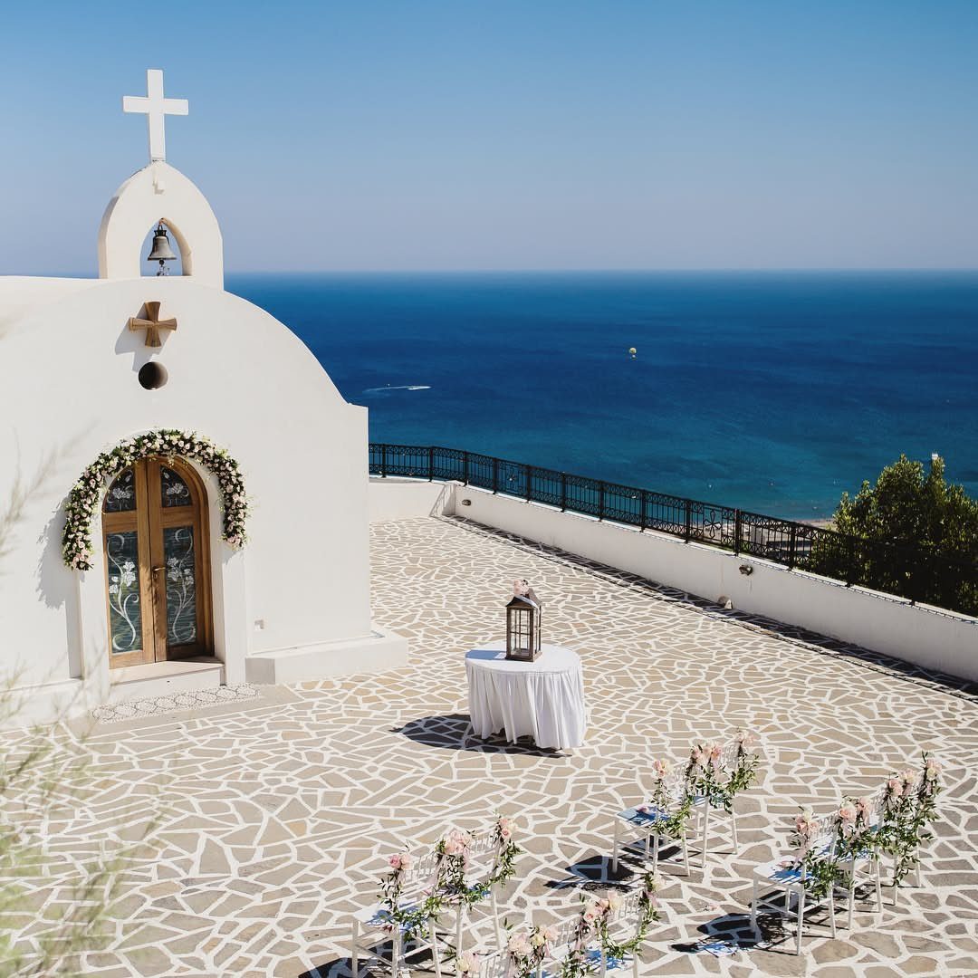 White Greek chapel overlooking the blue sea with wedding chairs and floral arrangements.
