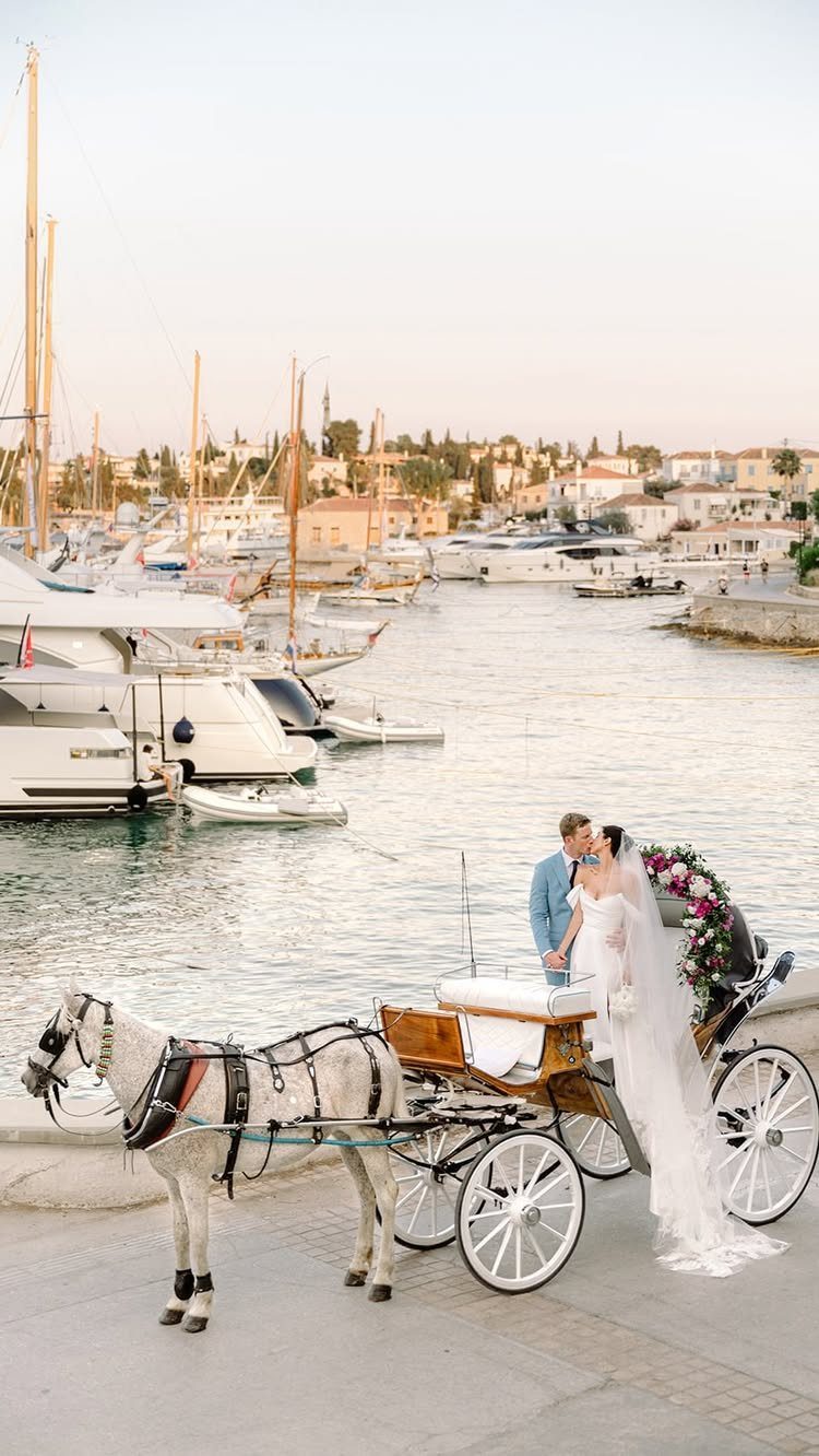 Bride and groom kissing by a horse-drawn carriage at a Greek harbor with yachts.