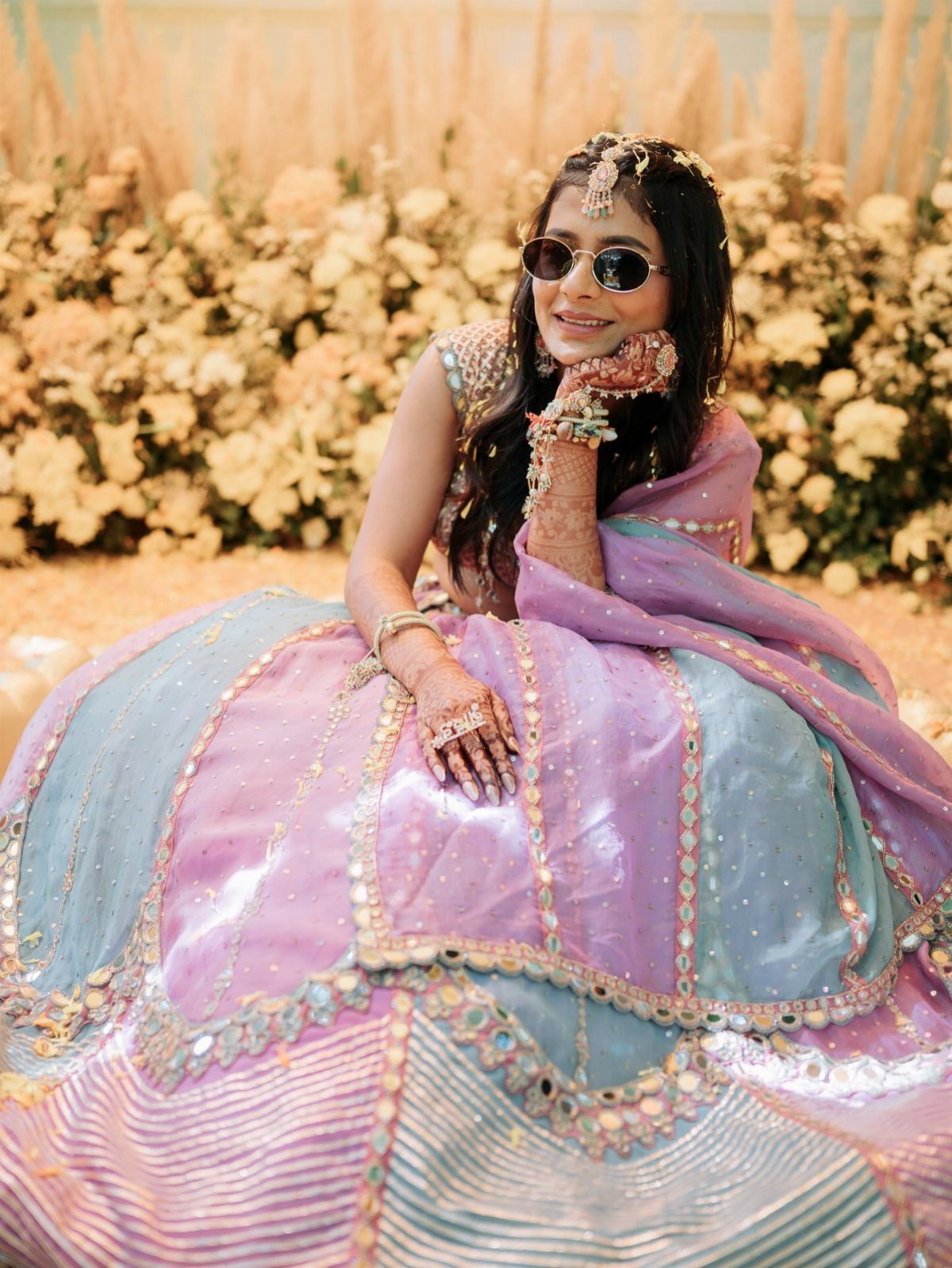 Woman in a colorful shimmering lehenga and sunglasses sitting before yellow flowers.