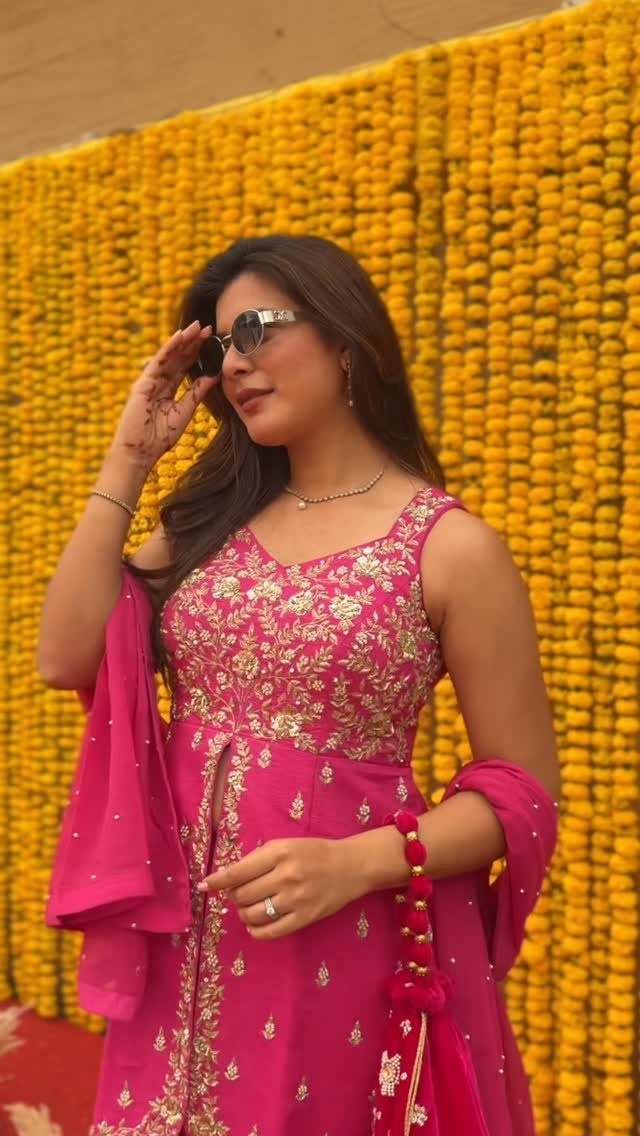 Woman in pink embroidered Indian outfit posing against a yellow marigold flower wall.