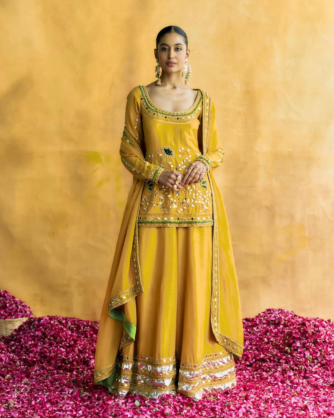 Woman in yellow mirror work kurta and wide leg trousers on pink petals.