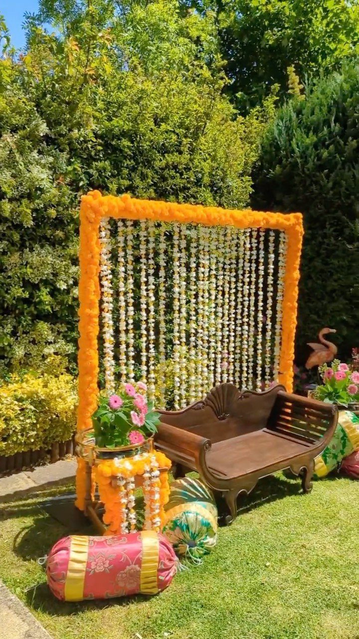13 Mehndi Decoration Ideas for Outdoor Parties 4 Wooden settee stage with orange and white floral backdrop on a green lawn.
