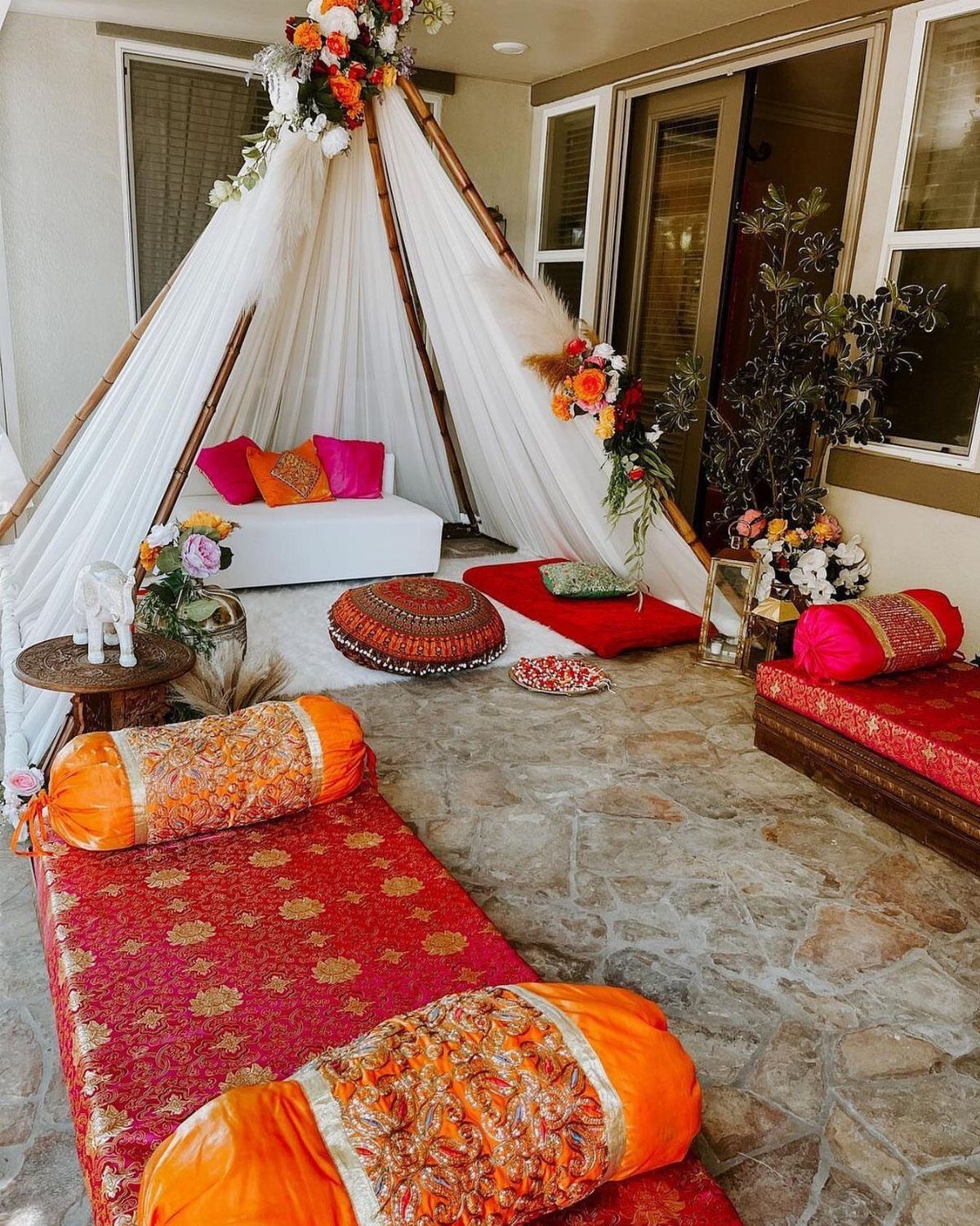 13 Mehndi Decoration Ideas for Outdoor Parties 10 White teepee tent with floral accents and colorful floor cushions on a stone patio