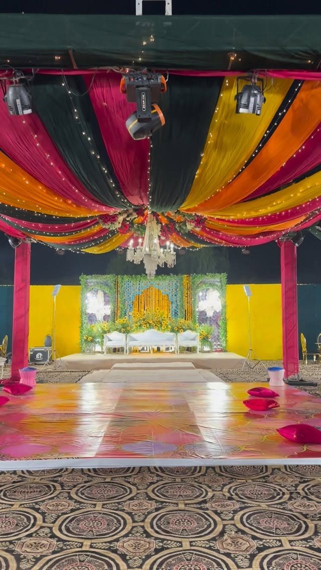 13 Mehndi Decoration Ideas for Outdoor Parties 11 Colorful draped fabric ceiling and floral stage setup for an outdoor mehndi celebration.