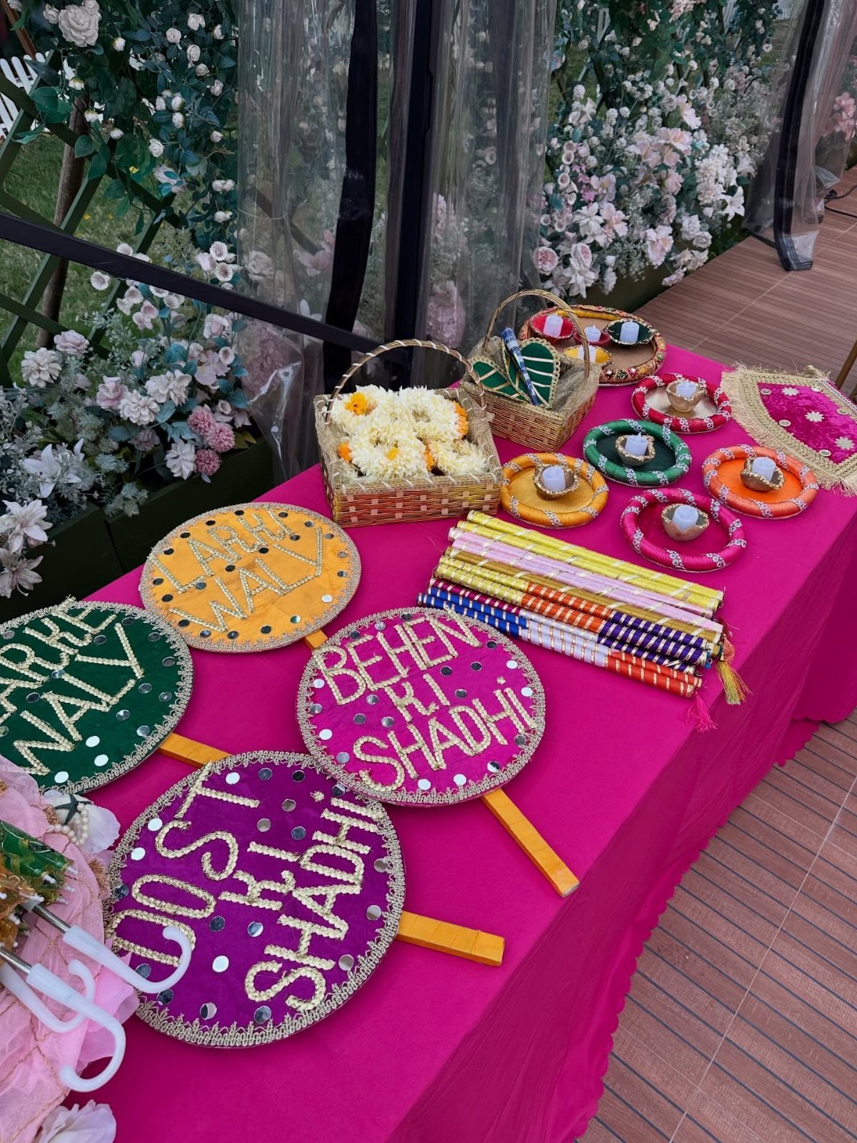13 Mehndi Decoration Ideas for Outdoor Parties 7 Colorful embroidered plaques and floral baskets on a pink table for a Mehndi event.