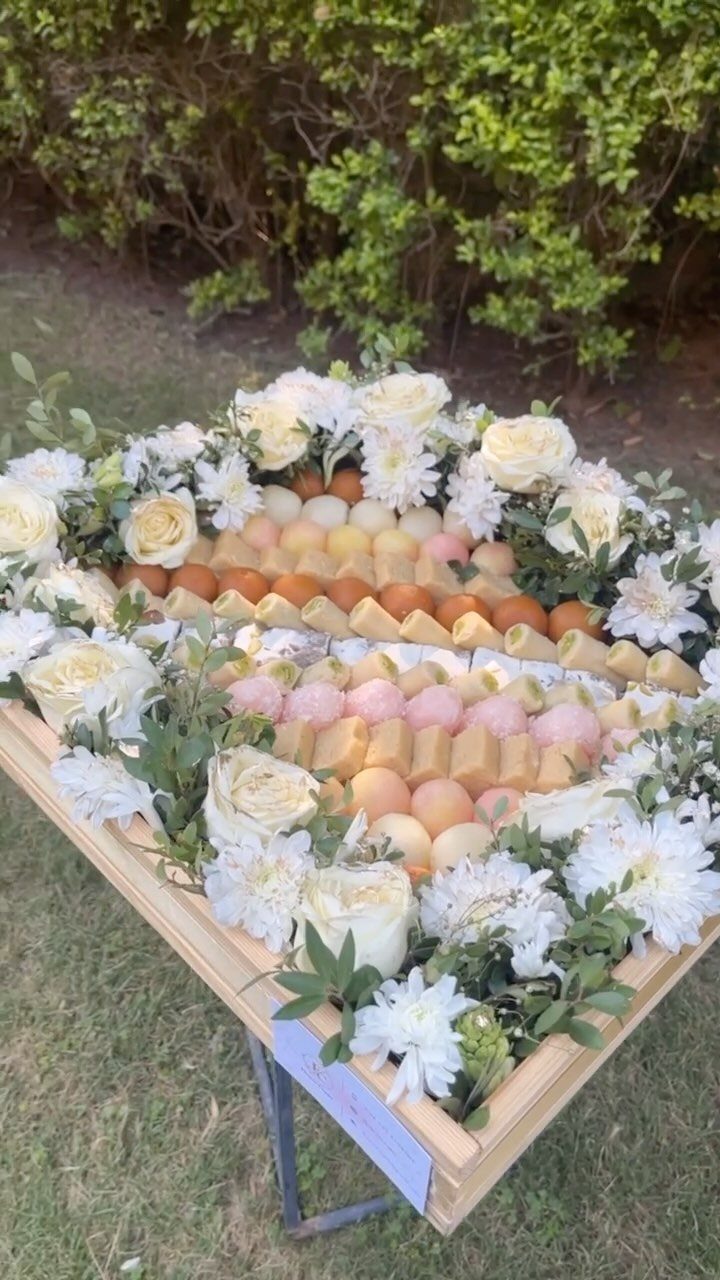 Wooden tray of colorful Indian sweets surrounded by white roses and chrysanthemums outdoors.