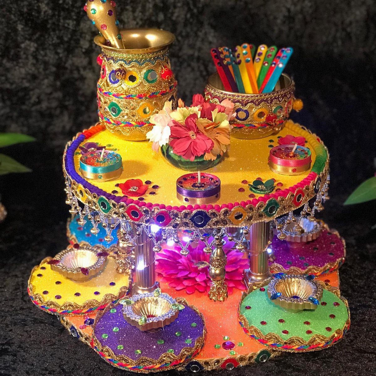 Multi-tiered colorful mirrored henna thali with gold pots, candles, and floral decorations.
