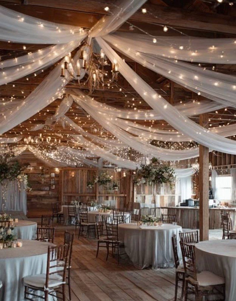 Rustic barn interior with white fabric draping, string lights, and set reception tables.
