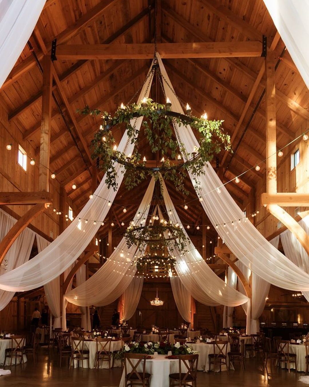 Wooden barn interior with white fabric draping, greenery chandeliers, and set reception tables.