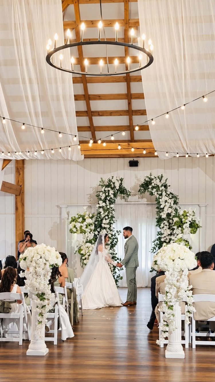 Bride and groom standing before a floral arch inside a modern white barn venue.