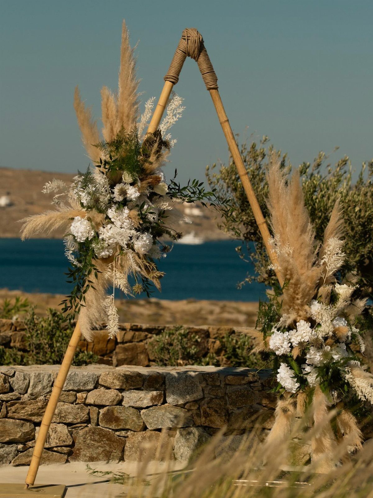 Bamboo triangle wedding arch with large white floral arrangements and fluffy pampas grass.