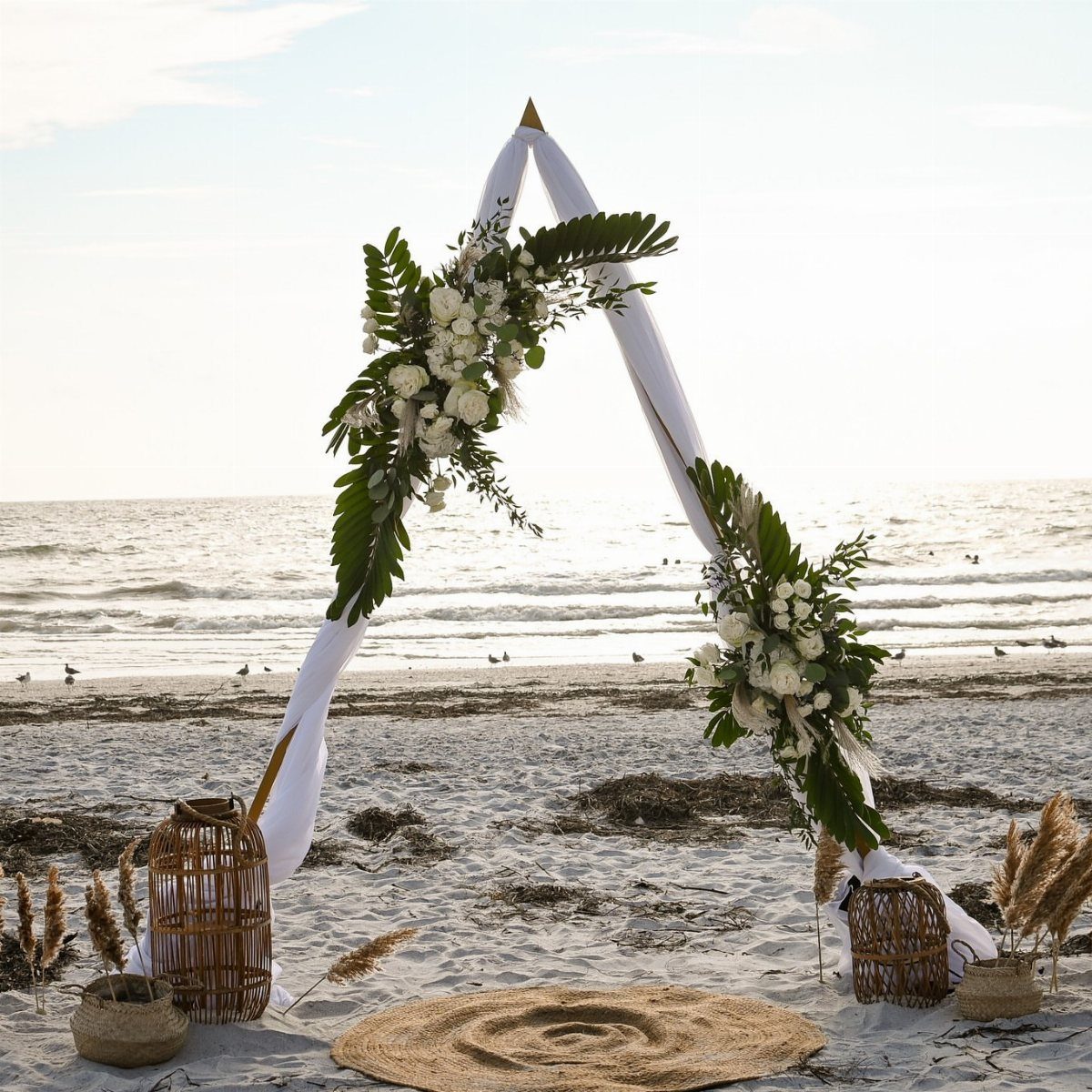White fabric triangle wedding arch with floral arrangements on a sandy beach