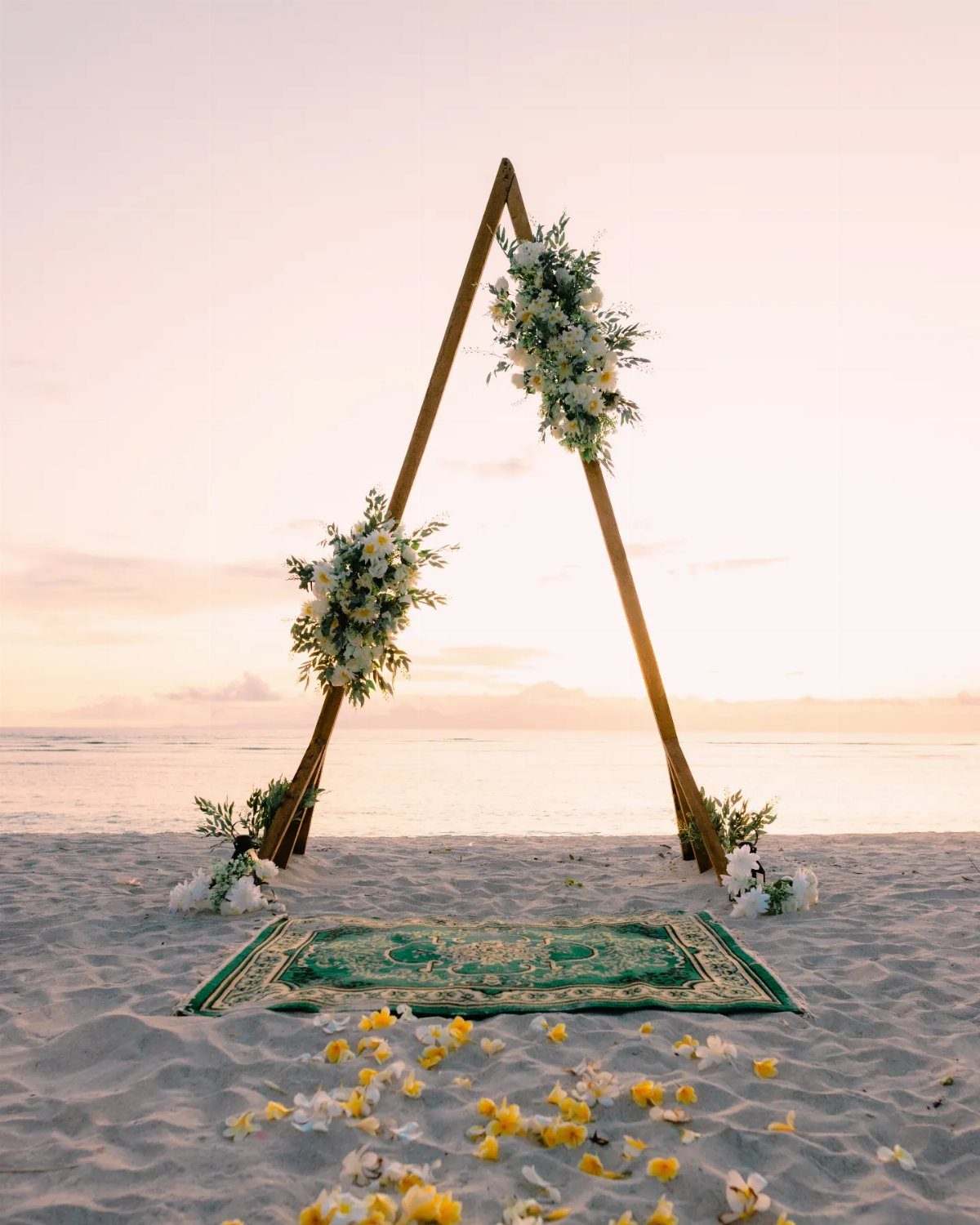 Wooden triangle wedding arch with white flowers on a beach with a rug.