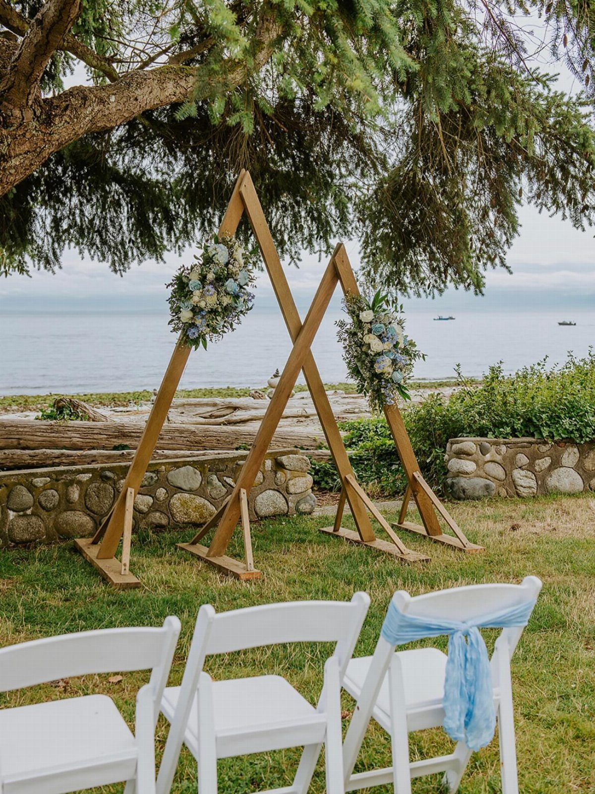 Wooden triangle wedding arch with blue and white flowers by the ocean shore.