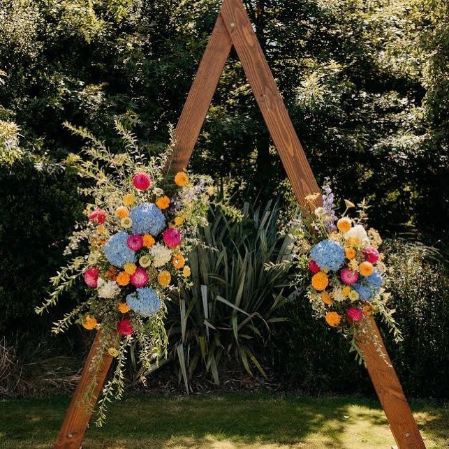 Wooden triangle wedding arch with colorful floral arrangements on a green lawn.