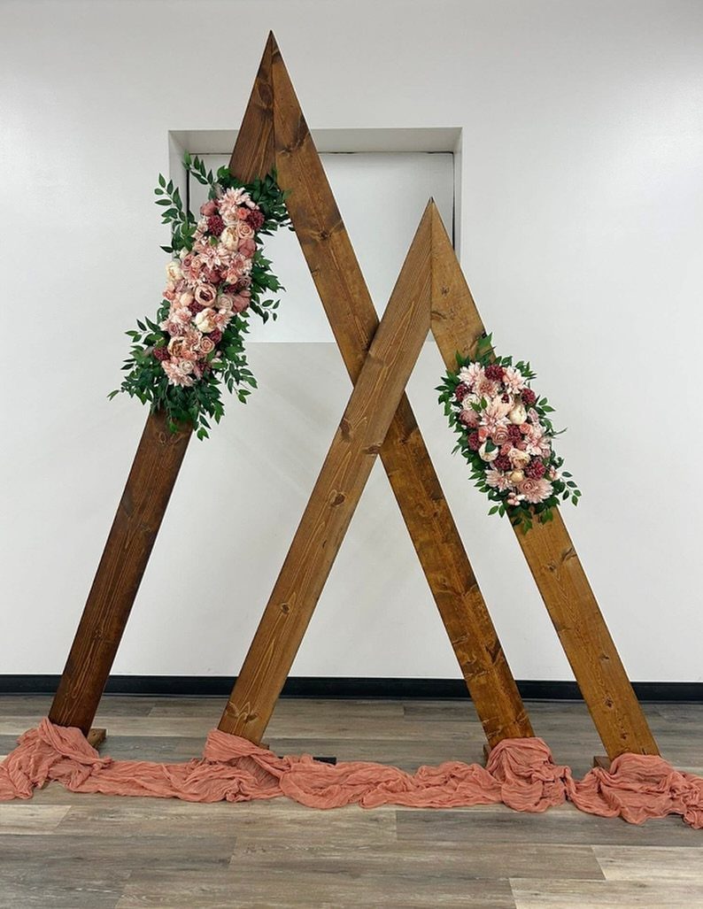 Wooden triangle wedding arch decorated with pink flowers and greenery against a white wall